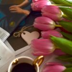 Pink tulips beside an art book, a small heart charm, and a cup of coffee