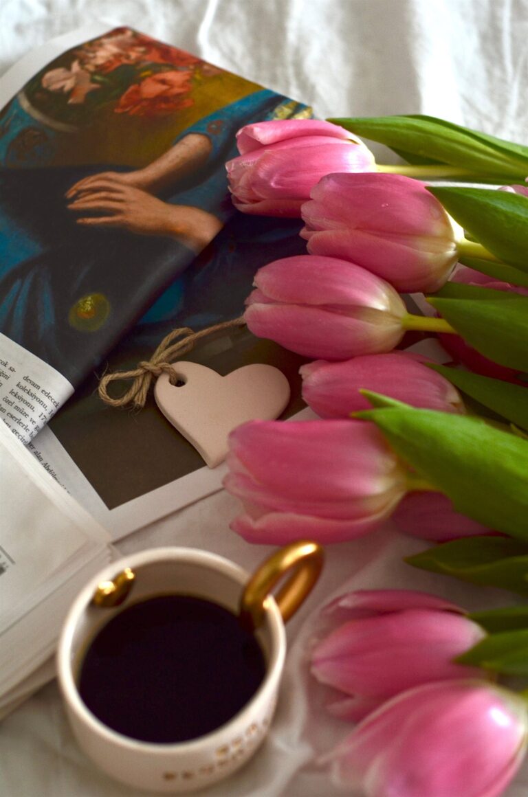 Pink tulips beside an art book, a small heart charm, and a cup of coffee