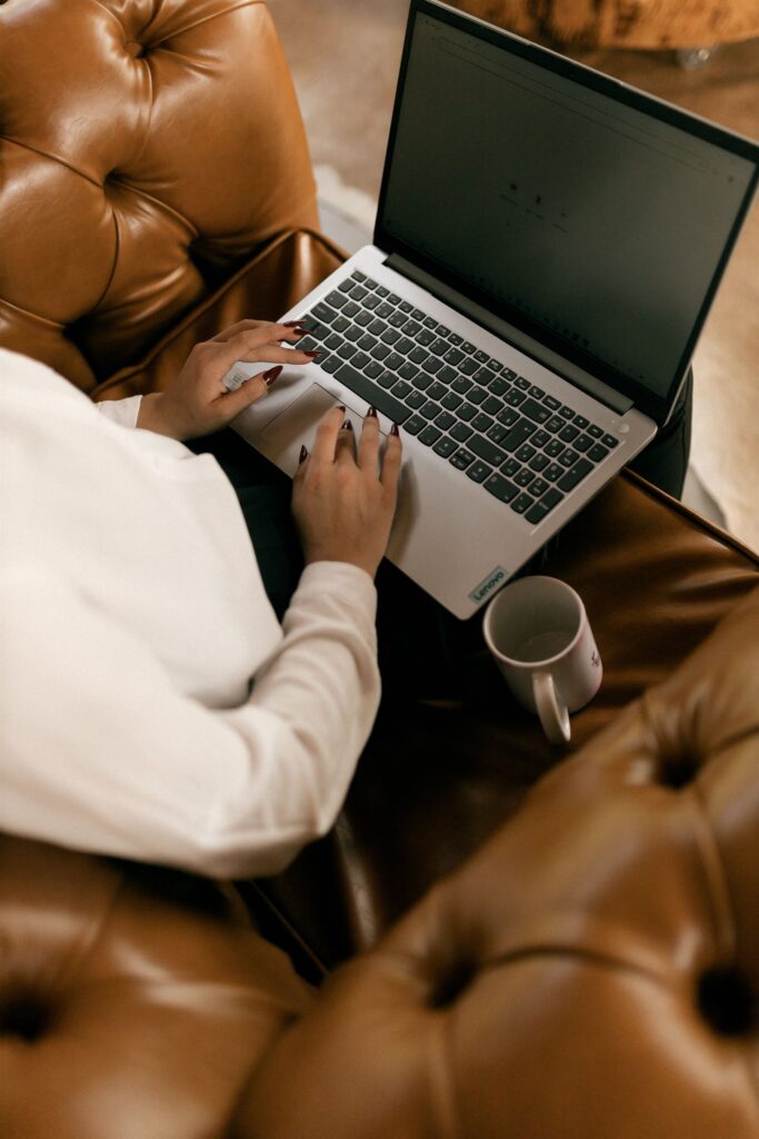 Woman writing on her laptop from a cozy leather armchair with a cup of tea